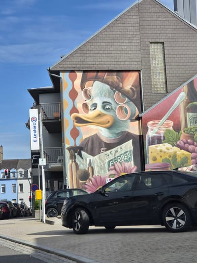 The image depicts a mural on the side of a building in Käerjeng, Luxembourg. The mural features a stylized duck-like character with curlers in its hair, reading a newspaper titled "NEWS FERBER." In front of the mural, several black cars are parked on a paved street. A tall, white sign for "E.Leclerc" is visible on the side of the building. The scene is set against a blue sky with some wispy clouds. The building itself has a grey, tiled roof and glass block windows. Traffic lights and street signs are also present in the background.