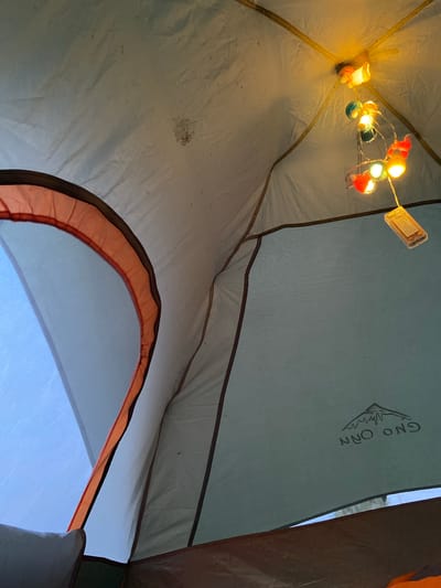 The image shows the interior of a tent with string lights overhead. The tent fabric is light-colored, and there are some dark spots on the ceiling. The string lights are multi-colored and illuminated. A small battery pack is attached to the lights. There is a mesh panel on the right side of the tent with a logo that appears to say "GNO ONN" and a mountain silhouette. The overall scene is dimly lit and appears to be at night or in low light conditions. The location is identified as Peshcherka, Russia.