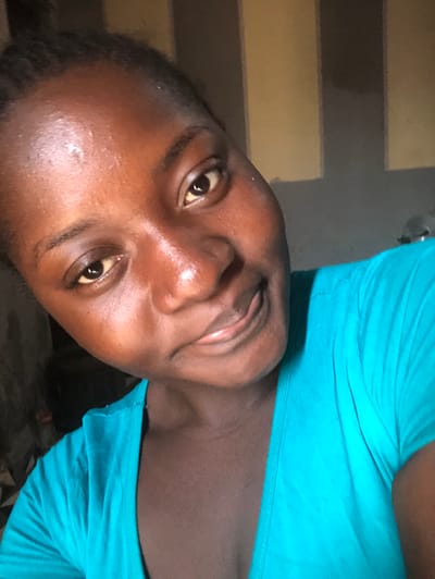 The image depicts a close-up, slightly upward-angled selfie of a young adult female with dark skin, located in Kujama, Nigeria. The subject directly faces the camera with a subtle, closed-mouth smile. Her dark hair is pulled back, and she wears a bright teal short-sleeved top. The background is an unfocused interior wall featuring vertical stripes or panels in alternating shades of light tan and dark gray. No other individuals, objects, or specific actions are discernible within the frame.