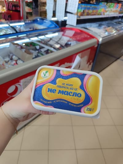 A hand holds a package of vegan butter substitute labeled "Не масло" which translates to "Not Butter." The package is yellow and pink with a vegan logo and the words "не можу повірити що це" which translates to "I can't believe it's" underneath the word "Not Butter." The package is 250 grams. The hand is holding the package in front of a blurred image of a refrigerated display case in a grocery store in Berehove, Ukraine (latitude: 48.197, longitude: 22.648). The floor is white tiled. The image is taken from a first-person perspective.