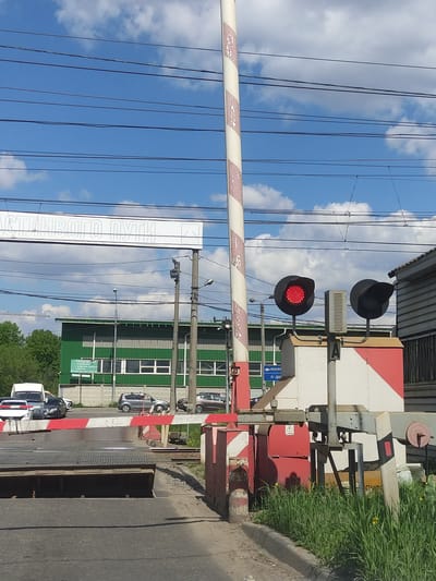 The image depicts a railway crossing in Elektrougli, Russia. The crossing gate is down, with red warning lights illuminated. A striped barrier blocks the road. A green and grey building is visible in the background. A sign with Russian text is located to the left. Several cars are waiting at the crossing. Overhead power lines are visible against a cloudy sky. The road surface is asphalt. Green grass and foliage are present on the right side of the image.