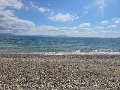 This is a wide-angle, eye-level shot of a pebbly beach in Astros, Greece. The foreground is dominated by the textured surface of the beach, composed of various sizes and colors of pebbles and sand. The middle ground features the gentle, clear blue waters of the sea, with small waves lapping at the shore. In the background, a distant mountain range is visible, silhouetted against a bright blue sky dotted with scattered white clouds. The sun is shining, creating a slight glare on the water and casting a light across the scene. There are no people or man-made objects visible in the image.