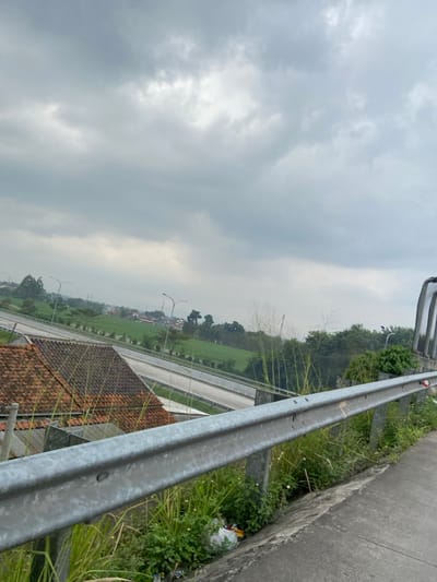 The image shows a view from a highway overpass in Kecamatan Margaasih, Indonesia. The overpass is made of concrete and has a metal guardrail. The highway is four lanes wide and has a concrete median strip. The highway is lined with trees and bushes. There are a few buildings in the distance. The sky is cloudy and overcast. The photograph was taken from the edge of the highway overpass, looking towards the city. There is a small amount of trash visible in the grass along the side of the highway. The view is from a high vantage point and gives a wide vista of the landscape. The lack of people in the photo suggests it was taken at a time when traffic is light.