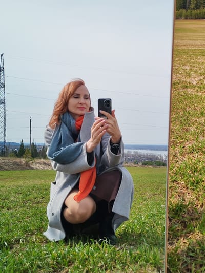 A woman with red hair is squatting in a grassy field, holding a smartphone to take a selfie in a mirror propped up beside her. She's wearing a grey coat with an orange and blue scarf. An electricity pylon is visible in the background under a blue sky. In the mirror reflection, a field and trees are visible as well as a body of water and a city with churches. This location appears to be in Nechkino, Russia.