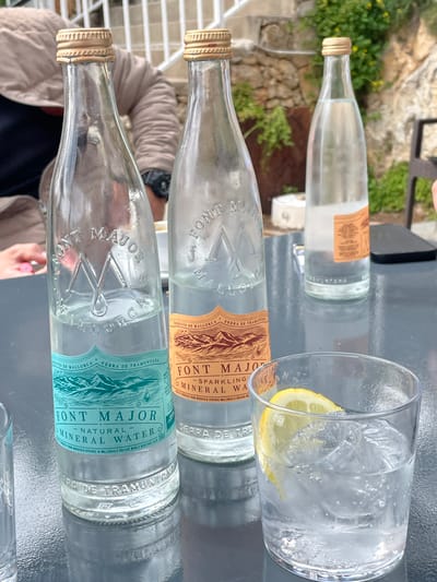 The image captures an outdoor table scene, likely at a cafe or restaurant in Porto Cristo, Mallorca, Spain. A dark grey, textured tabletop dominates the foreground, holding three glass bottles of "FONT MAJOR" mineral water. The leftmost bottle, labeled for "Natural Mineral Water" with a teal mountain design, is approximately half full. Adjacent to it, a second bottle, labeled for "Sparkling Mineral Water" with an orange mountain design, is also partially full. A third, almost empty "FONT MAJOR" bottle stands further right. In the immediate foreground, a clear glass tumbler contains ice cubes, a clear liquid, and a yellow lemon slice. On the right side of the table, a dark smartphone is partially visible. In the upper left corner, a person's arm, clad in a light beige jacket, is visible, with a hand positioned near a pink object, possibly a straw. The background features a light-colored stone wall adorned with green foliage and a white railing structure.