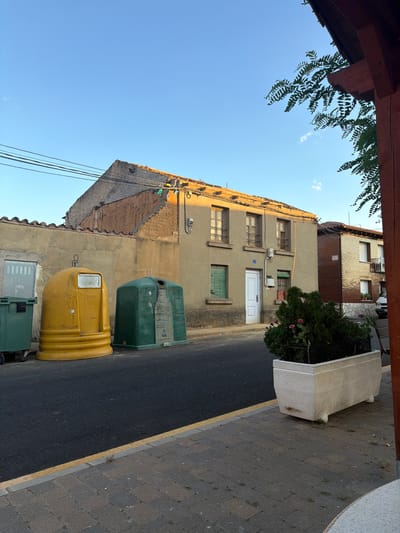 The image depicts a street scene in Arcahueja, Spain, with a focus on two large, colorful recycling bins on the side of the road. To the left, a tall green recycling bin is partially visible. Next to it stands a large, rounded yellow bin, followed by a dark green rectangular bin. Behind the bins, a weathered, light-colored wall runs along the side of a building. This building has a sloped, damaged roof with exposed rafters and some tiles missing. To the right of this building is a narrower brick building with several windows and a balcony railing on the upper floor. The street itself is paved with asphalt, marked by a yellow line along the curb. The sidewalk is made of grey brick pavers. In the foreground, a white rectangular planter box contains green bushes and a few pink flowers. The sky above is clear blue with a few wispy clouds. Sunlight casts warm tones on the buildings, suggesting it is late afternoon or early evening. No people are visible in the frame.