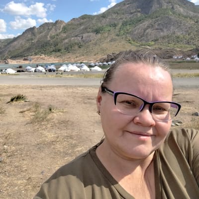 A middle-aged woman with light brown hair and glasses is taking a selfie in an outdoor setting. She is wearing a dark green t-shirt. Behind her, a road is visible with several white yurts set up in rows, suggesting a tourist area or settlement. Cars are parked near the yurts. In the background, a large, rocky mountain looms under a partly cloudy blue sky. The foreground consists of dry, dusty ground with sparse vegetation. The location is identified as Torayghyrov, Kazakhstan.