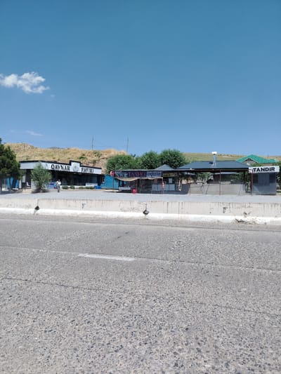 The image captures a sunny day in a rural or semi-rural setting, likely in Qaynar MFY, Uzbekistan. The scene features a collection of roadside establishments, including a building with "QAYNAR PATIRI" visible on its facade, suggesting it's a restaurant or food service establishment. Adjacent to it, another structure bears the name "TANDIR," and further down, "TANHO MINI MARKET." The establishments appear to be open-air or have open sides, with covered roofs.
In front of the buildings, a low concrete barrier separates them from a paved road. A few trees are scattered around the area, and a grassy, undulating hill forms the backdrop. A solitary person is visible walking near the "QAYNAR PATIRI" building. The sky is a clear blue with a single, small white cloud. The overall impression is one of a commercial area catering to travelers or locals, operating in warm, daytime weather. The presence of the "TANDIR" sign indicates the likely availability of traditional baked goods.