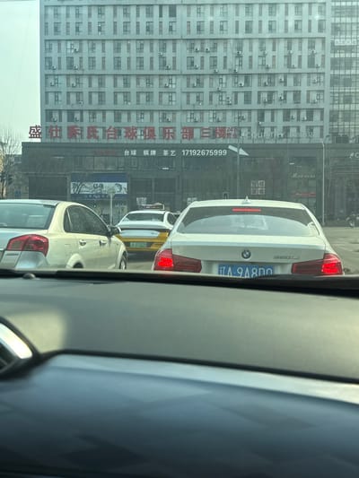 The image depicts a daytime urban street scene captured from inside a vehicle, with the dashboard partially visible in the foreground. The location is Shenhe District, Shenyang, Liaoning Province, specifically near No. 1 Chaoyangli, Chaoyang Street.

Three vehicles are prominently featured: a white BMW 320Li sedan directly in front of the viewer's vehicle, displaying illuminated red brake lights and a partial license plate "辽 A 9A8D0"; a light-colored sedan (silver or champagne) partially visible to the BMW's left; and a white and yellow taxi with license plate "辽 A D75G0" positioned further back in a middle lane. All vehicles appear stationary, indicating traffic congestion or a stop at a traffic signal.

In the background, a large, multi-story grey building with a repetitive grid of rectangular windows dominates the upper frame. Numerous external air conditioning units are affixed below many of these windows. The lower, darker commercial section of this building displays large red Chinese characters identifying "盛社新氏台球俱乐部三楼" (Sheng She Xin Billiards Club Third Floor). Below this, smaller black text advertises "Billiards, Chess & Cards, Tea Art" and a phone number (17192675999). Partial signage indicating "24H" and "COUNTRY" is visible to the right of the billiards club name. To the far right, a segment of a modern glass-paneled building is also visible.

The sky is bright but appears somewhat hazy. A green traffic light is faintly visible in the distance to the left. No individuals are clearly discernible in the scene.