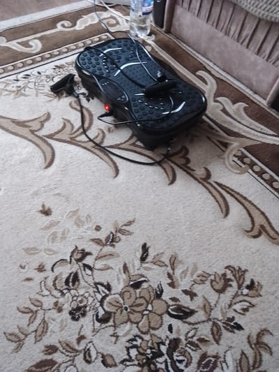 The image shows a black vibrating fitness platform with a red light indicator, placed on a carpet. The platform has a textured surface with small bumps and white lines. A black cable is connected to the platform, and a small black handle or strap is attached to the left side. To the left of the platform, the edge of a carpet with a brown and beige floral pattern is visible. In the background, a portion of a sofa with a pleated fabric in a light brown or beige color is present, along with a partially visible plastic bottle. The scene appears to be indoors, possibly in a living room, with the lighting suggesting daytime. No people are visible in the image. The setting is likely Shakhtarsk, Ukraine, as indicated.