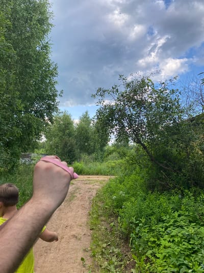 The image depicts a hand holding a pink leash, with a child's head visible in the lower right corner, suggesting they are walking together. They are on a dirt path surrounded by lush green foliage and trees. The sky above is a mix of blue and gray clouds, indicating potentially mixed weather. The sunlight filtering through the trees suggests it is daytime. The overall scene evokes a sense of a casual outdoor activity, possibly a walk in a park or a nature trail in Novyi Ural, Russia. There is no visible text in the image.