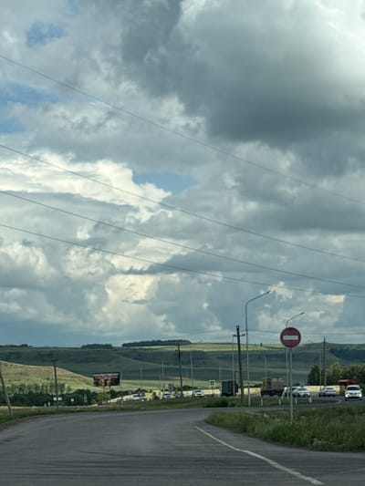 The image depicts a road scene on an overcast day, likely in or near Kendektamak, Russia. The foreground is dominated by a paved road curving to the left, with white lane markings visible. To the right of the road, overgrown grass and vegetation are present. In the middle ground, a two-lane road is visible with several vehicles, including cars and trucks, traveling in both directions. A traffic sign indicating "No Entry" is prominently displayed on a pole on the right side of the road. Further in the background, rolling green hills stretch across the landscape, dotted with trees and some scattered buildings. A large billboard is visible on the left side of the middle ground, advertising what appears to be an event or product. Overhead, a complex network of power lines crisscrosses the sky. The sky itself is filled with dramatic, dark gray clouds, suggesting imminent rain or a recent storm. There are no people clearly visible in the image.