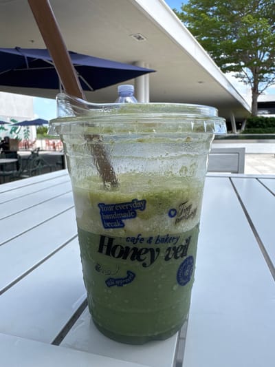 An outdoor scene in Hallandale Beach, Florida, United States of America, shows a close-up of a green, blended beverage in a clear plastic cup with a lid and a wooden straw. The cup has blue text on it that reads "Your everyday handmade break," "Tiny treat shop," "cafe & bakery," and "Honey Veil." There is also a small bee illustration with the text "uki approved." The cup is condensation-covered and sits on a white slatted table. In the background, there are blue umbrellas, a tree, and a modern building. The lighting suggests it is daytime.