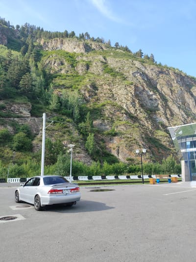 A silver sedan is parked in a paved lot at the base of a rocky, tree-covered mountain. The car is facing away from the viewer and is parked near a white utility pole and a streetlamp. In the background, a modern building with glass windows is visible, partially obscured by trees and shrubbery. The sky is clear and blue. The scene appears to be located in a scenic area, possibly a park or a rest stop, near Cheremushki, Russia. There are no people visible in the image.