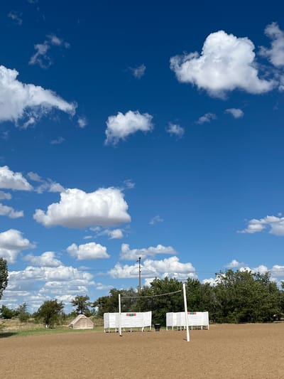 This image depicts an outdoor scene, likely a sandy volleyball court, under a bright blue sky dotted with fluffy white clouds. In the foreground, the court surface is smooth and brown. A volleyball net is strung between two white posts. On either side of the net are white structures with colorful markers, possibly scoreboards or signage. In the background, a variety of trees form a green backdrop. A beige tent is visible to the left of the court, and a utility pole with lights stands to the right. The overall environment suggests a recreational area in a somewhat rural or open setting. There are no people or animals visible in the image. The location is specified as Gres-2, Kazakhstan.