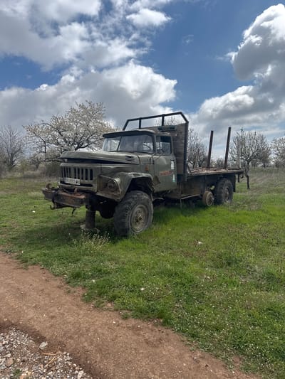 The image shows an abandoned, damaged, olive-green truck parked in a grassy field near a dirt road, likely in Gorna Grashtitsa, Bulgaria. The truck has a flatbed, appears to be a GAZ-66, and is missing a front wheel. The front of the truck is heavily damaged, with a mangled bumper and grill. A license plate is visible with illegible characters. In the background are flowering trees and a partially cloudy blue sky. The field is covered in green grass with small white flowers. A dirt road borders the grass. There are no people visible.