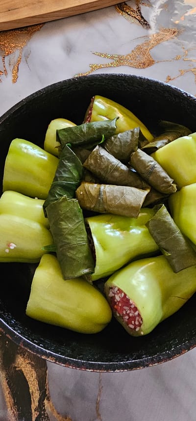 A black bowl contains a dish of stuffed green peppers and grape leaves. The green peppers are cut lengthwise and filled with a mixture of ground meat and rice. Some of the peppers are whole, while others are cut in half. The grape leaves are stuffed with a similar mixture and rolled into small cylinders. The bowl is placed on a marble-patterned surface with gold accents. The scene is lit from above, casting soft shadows. There are no people visible in the image. The location is the village of Shyngyrly rural district, Kazakhstan.