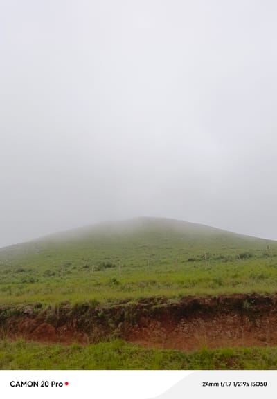 The image shows a grassy hillside shrouded in fog, a common sight in Nguroje, Nigeria, a town known for its cool climate and misty mornings. The foreground features a dirt embankment with sparse vegetation, leading up to the gently sloping hill. A few wooden fence posts are visible on the hillside. The sky is completely obscured by thick white fog, creating a serene and somewhat mysterious atmosphere. The bottom of the image displays text indicating the camera model, "CAMON 20 Pro," and photographic settings: "24mm f/1.7 1/219s ISO50." There are no people or animals visible in the scene.
