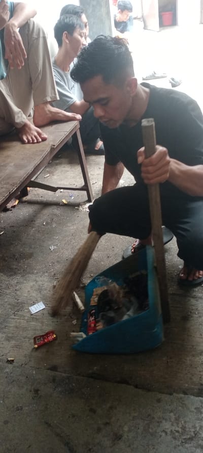 A male individual in Cilampeni, Indonesia, is captured in a crouched position, actively sweeping debris into a blue plastic dustpan on a concrete floor. He wears a black short-sleeved shirt, dark pants, and black flip-flops, holding a natural-fiber broom in his right hand and the dustpan in his left. The dustpan contains various wrappers and refuse, while additional discarded items, including a red wrapper and a piece of paper, lie on the floor nearby. To the left, three other males are partially visible. One is seated on a low, unpainted wooden bench, with bare feet resting on its surface. Another, wearing a grey t-shirt, is behind him. A third male's head is seen further back. The background features a light-colored wall and an open doorway revealing an interior with a red bucket. Scattered footwear is visible on the concrete floor in the background.