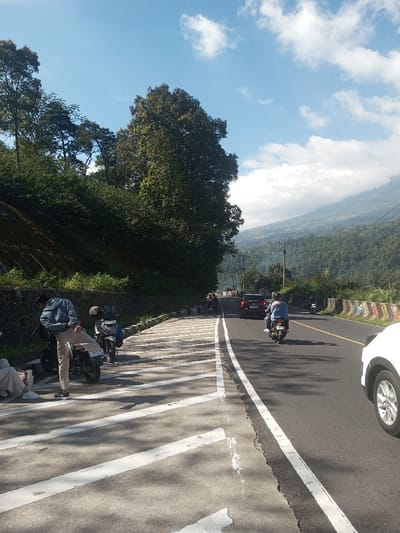 The image captures a sunny daytime scene along a two-lane asphalt road in Ka Thaung (Upper), Myanmar, with a clear blue sky partially obscured by white clouds. On the left side of the road, a steep, densely vegetated hillside rises, bordered by a low, dark stone wall. Adjacent to the wall is a wider paved shoulder featuring prominent diagonal white markings.
On this left shoulder, two individuals and two motorcycles are present. One person, wearing a light blue denim jacket and light-colored pants, stands with their back to the camera, leaning against a parked black motorcycle. Another person, dressed in light-colored clothing, is partially visible seated further to the left foreground. A second dark-colored motorcycle is parked behind the first.
The asphalt road has a solid white line separating traffic lanes and a white line marking the right edge of the road. Vehicles are traveling away from the camera. In the right lane, a person wearing a blue top and a helmet is riding a blue motorcycle. Further down the road in the same lane, a dark-colored sedan or SUV is visible. On the far right foreground, the front wheel and lower body of a white vehicle are partially visible.
In the mid-distance on the right, utility poles and lines are visible, and the road curves slightly. A section of a roadside barrier adorned with colorful graffiti is present on the right side. Beyond the road and extending into the background, forested mountains rise, partially obscured by clouds at their peaks.