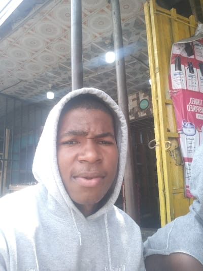 The image displays a close-up scene in Igwuruta, Nigeria, featuring a young Black male and partial view of a second individual, situated in what appears to be a semi-enclosed commercial or residential area. The primary subject, positioned on the left two-thirds of the frame, is a young male wearing a grey hooded sweatshirt with the hood up. He looks directly at the camera, exhibiting a slight furrow in his brow and a closed mouth. To the bottom right, the right shoulder and upper arm of a second person, also in a grey hooded sweatshirt, are visible.
Overhead, a ceiling or awning composed of light-colored tiles with a repeating white and orange/brown circular pattern is visible. Several white, circular light fixtures are recessed into this ceiling. Two dark, vertical poles stand behind the primary subject. To the right of the primary subject, a weathered yellow metallic structure, consistent with a door frame or gate post, is present. Attached to this yellow structure is a red and white poster, advertising electronic devices, with text such as "TUNED INSPIRED" and "AIRPODS" appearing partially inverted. In the background, to the left of the yellow structure, a darker interior space is visible, containing a circular light green sign on a wall. The scene is brightly illuminated by direct sunlight, casting strong highlights on the primary subject and generating a lens flare near the top of the central pole.