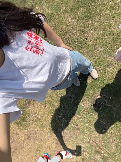 The image shows a first-person perspective of a person standing on grass, presumably in El Oasis Valsequillo, Mexico, given the prompt. The person is wearing light blue jeans, a white t-shirt with red text, and beige shoes. The text on the t-shirt reads "Living Ya Life in SPRING! COOL, SUNNY". The person's hair is dark and visible on the lower left. There are shadows visible on the grass, including the shadow of the person taking the photo and at least one other person wearing novelty shoes, red, white, and blue, standing to the right. The grass appears to be short and dry in places.