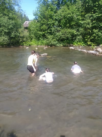 The image shows three people in a shallow river. Two are partially submerged, and one is standing in the water. The person on the left is wearing a yellow top and black shorts, with their back to the camera. The person in the middle is wearing a white t-shirt and appears to be playing with the person in front of them. The person on the right is wearing a white t-shirt and is facing away from the camera, with only their head and shoulders visible above the water. The river has a rocky bottom and is surrounded by lush green trees. The sun is shining, suggesting it is daytime and the weather is likely warm. The scene appears to be in a natural, outdoor setting. The image is captured from a high angle, looking down at the river.
