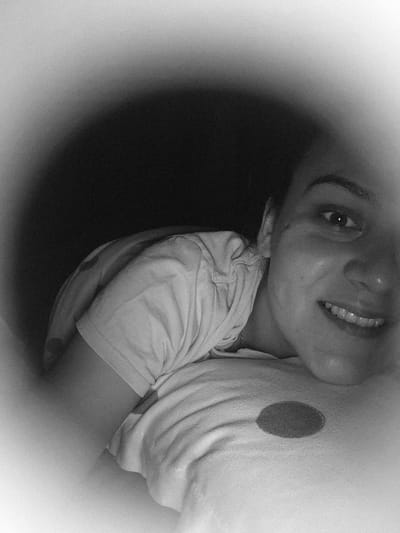 A black and white, heavily vignetted photograph shows a person, likely female, in a close-up, horizontal orientation. The image is characterized by significant digital noise and a bright, almost white, circular border transitioning to a dark, unlit background around the central subject. The individual's right side of the face (viewer's right) is partially visible and illuminated, revealing a smiling expression with both upper and lower teeth exposed. The right eye is open, looking directly towards the viewer. Dark eyebrows are present above the eye, and the nose and mouth are clearly defined. The person's ear is partially visible on the left side of the face, near the neckline.
The individual is wearing a light-colored, short-sleeved t-shirt, which shows distinct fabric texture and folds across the shoulder and chest area, consistent with lying on one's side. A dark, oval-shaped marking or patch is visible on the t-shirt, situated on the lower left portion of the visible garment. The background surrounding the subject is uniformly dark and devoid of discernible features. The geographic location, specifically the city of Iskrets, Bulgaria, cannot be determined from the visual content of this image.