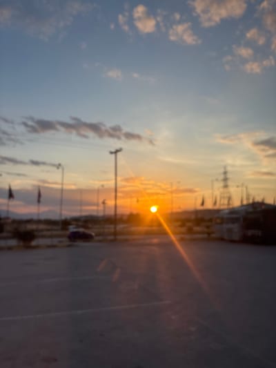 The image captures an outdoor scene at sunset, rendered with significant blur. The sky, occupying the upper two-thirds of the frame, transitions from a pale blue with scattered, soft-edged cumulus clouds at the zenith to an intense gradient of orange and yellow near the horizon. The setting sun, a brilliant white-yellow point of light, is positioned in the mid-ground, slightly to the right of the vertical center, emitting a pronounced, diagonal lens flare extending downwards and to the left.
The foreground features a dark gray asphalt surface, characteristic of a parking lot or road, with faint light-colored markings visible in several areas. The mid-ground contains various elements: a dark-colored passenger vehicle is discernable on the left side, appearing stationary. Multiple utility poles and streetlights are distributed across this section. Several dark, rectangular flags are mounted on poles in the left-mid ground, their details obscured by blur. On the right, a prominent, dark-colored power transmission pylon with a lattice structure is visible, alongside the indistinct shapes of large vehicles, possibly commercial trucks or buses, partially obscured by structures. The background presents a hazy, dark silhouette of distant mountains or hills against the colorful horizon.
No individuals are clearly identifiable within the frame. The overall image is heavily out of focus, contributing to a soft, almost painterly effect, with light and color being dominant elements. This scene, featuring infrastructure, vehicles, and a mountainous backdrop under a sunset sky, aligns with locations often found in the Municipal Unit of Lamia, Greece, known for its mix of urban development and surrounding natural landscapes.