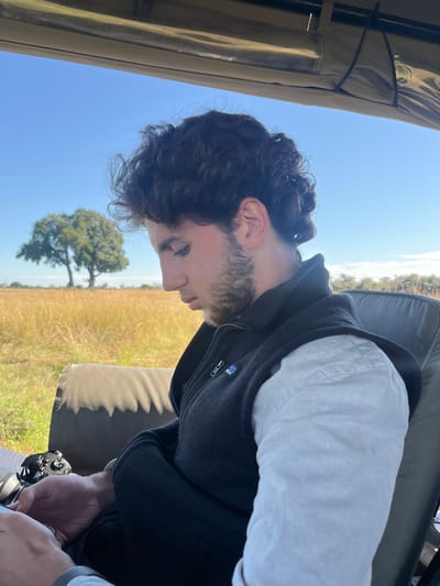 A man with dark curly hair, a beard, and wearing a black Patagonia fleece vest over a white shirt is seated in what appears to be an open-top safari vehicle. He is holding a black camera. The setting is outdoors, with a field of tall grass and a single tree visible in the background under a clear blue sky. The vehicle has a tan canvas roof. It is assumed the image was taken in Kakoro, Botswana.