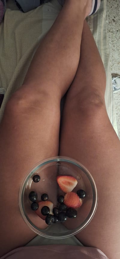 A high-angle, close-up shot captures a person's legs, likely bare, resting in a relaxed position. The legs are seen from the thighs down to the calves, with a light-colored blanket or sheet partially visible beneath them. A clear glass bowl containing a mix of strawberries and blueberries rests on one of the upper legs. The strawberries are cut into halves, and there are several dark blueberries scattered within the bowl. A small portion of a foot wearing a white sock is visible in the bottom-left corner of the frame. The background shows a light-colored, textured surface, possibly a wall or floor. The scene depicts a moment of leisure, possibly enjoying a healthy snack while relaxing indoors in Santa Cruz De Tenerife, Spain. No visible text is present in the image.