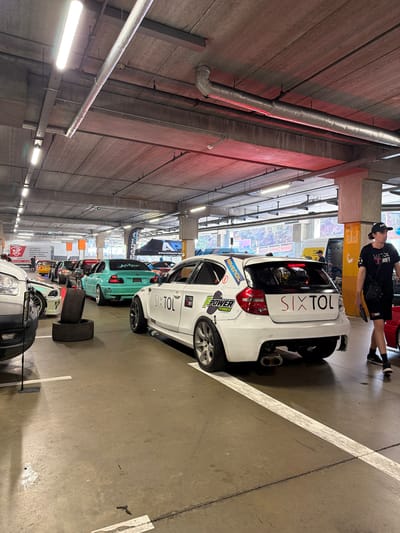 In a dimly lit, multi-level parking garage, likely in Liberec, Czechia, a car show or event is underway. The concrete ceiling is crisscrossed with pipes and illuminated by fluorescent lights and red ambient lighting. In the foreground, a white modified hatchback, likely a BMW 1 Series, is parked. It features custom decals, including "SIX TOL" in large red lettering on the rear, and "POWER TIRES" and a dragon graphic on the side. Two used tires are stacked in front of it. Behind this car, a line of other modified cars is visible, including a bright teal sedan with "KAŠPAR" on its rear windshield, and several other vehicles in various colors. A person in a black t-shirt and shorts, wearing a baseball cap, walks away from the white hatchback, carrying a tablet. In the background, tents and banners suggest an organized event. The overall scene depicts a gathering focused on automotive customization and performance.