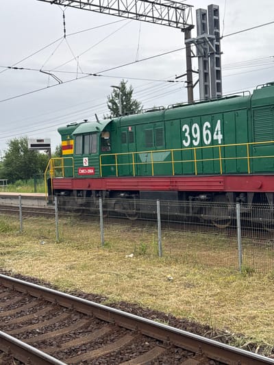 This image shows a green train engine with the number "3964" printed in white on its side. The train is on a railway track, with grass and a fence visible to the right. Overhead power lines and a signal gantry are present in the background, suggesting an active railway environment. The sky is overcast, indicating a cloudy day. The text "CHЕЗ-3964" is visible on the side of the train, near the front. The overall scene depicts a train at a station or on a track, possibly in Olaine, Latvia, given the location context. There are no people visible in the image. The lighting suggests it is daytime, but the overcast sky prevents a clear indication of the time.