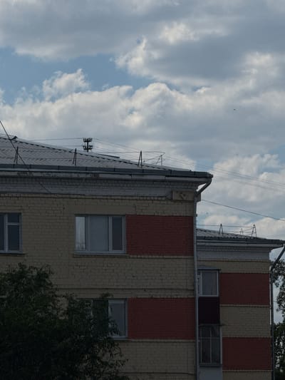 The image shows a close-up of the upper floors of two adjacent brick buildings, likely apartment blocks, in Zhovte, Ukraine. The building in the foreground has a facade of yellow brick with horizontal bands of red brick. It features several windows with light-colored frames. A grey metal roof with visible antenna structures and wires is visible at the top of the building. The building in the background, partially visible, shares a similar architectural style with yellow and red brickwork and a grey metal roof. The sky above is partly cloudy with white cumulus clouds against a light blue sky. A small dark object, possibly a bird or insect, is visible in the upper right portion of the sky. Trees with dark green foliage are visible in the lower left and right edges of the frame. The lighting suggests it is daytime, with the sun possibly behind the clouds or at an angle that creates soft shadows.