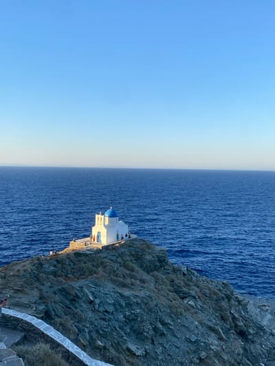 A vertical photograph captures a whitewashed Greek Orthodox church with a blue dome and twin bell towers, perched atop a rocky promontory that juts into the deep blue Aegean Sea. The church, known as the Church of Seven Martyrs (Epta Martyres), is located on the coast of Sifnos, Greece.
The church structure is pristine white, contrasting sharply with its vibrant blue dome. A small number of individuals, approximately 10-15, are visible on the paved area surrounding the church and on the connecting pathway, their specific actions indistinguishable due to distance.
The rocky promontory consists of dark, rugged stone with sparse, low-lying dry vegetation. A winding stone path with whitewashed accents is visible in the lower left foreground, leading down from an unseen vantage point towards the church. The sea, a consistent deep blue with subtle surface ripples, extends to the horizon where it meets a clear, light blue sky. Faint landmasses are discernible on the distant horizon line. The scene is illuminated by natural sunlight, casting a warm glow on the church and producing clear shadows on the rocky terrain, indicative of late afternoon or early morning light.