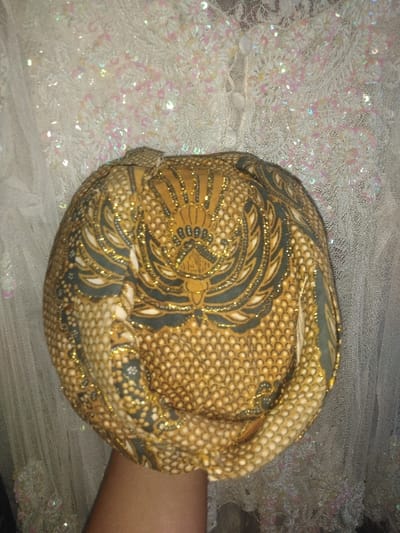 Location: Jehonville, Belgium. The image displays a traditional Indonesian head covering, likely a blangkon, positioned centrally and held upright. The fabric of the head covering is primarily golden-tan with intricate patterns in dark green-black and white. A prominent stylized bird motif, possibly a Garuda, with fanned wings and a decorative head, is centered at the top. The surrounding fabric features detailed scale-like, dot, and scroll patterns, all highlighted with golden glitter. The lower portion of a hand with brown skin is visible beneath the head covering, supporting it from below. The background consists of a white, sheer fabric adorned with numerous iridescent sequins or beads that form floral or circular patterns. This background fabric appears to be part of a garment or decorative textile.