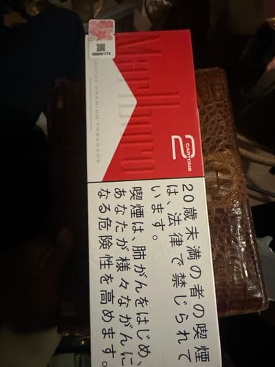 A pack of Marlboro cigarettes labeled "2 CARTONS" is resting on top of a brown leather wallet. The side of the pack displays a health warning in Japanese, indicating that smoking is prohibited for individuals under 20 years of age and increases the risk of various cancers. A serial number "000000177A" is visible on the side of the pack. The setting appears to be indoors, with a person wearing a blue shirt partially visible in the lower portion of the image. The exact location in Montreuil, France, is not identifiable from the provided visual information.