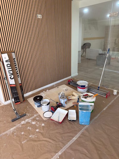This image depicts a room undergoing renovation or construction, specifically located in Gondomar, Portugal. The scene is characterized by unfinished work and tools commonly associated with painting and tiling.
The walls are partially covered with vertical wooden slats, suggesting a modern design element. On the left side, a large cardboard box with the brand name "SKUDA" is propped against the slatted wall. Next to it, a silver pole with a brush attachment, likely a vacuum cleaner extension, lies on the floor.
The floor is covered with brown protective sheeting, splattered with white paint in various locations, indicating active painting work. Several paint cans are present, including a large white bucket with blue and red accents, another white can with a blue lid, and a smaller black can. Paint rollers, paint trays, and brushes are scattered around, some on a wooden board. A tile cutter is also visible on the floor.
In the background, through a glass partition, another part of the building is visible. This area appears to be a hallway or a larger open space with flooring and some furniture. There are also signs of ongoing work in that area, including what appears to be a person in the distance and a large grey bin. A long-handled mop or squeegee leans against the glass partition.
The overall impression is one of a work in progress, with the necessary tools and materials laid out for painting and possibly tiling. The lighting suggests it is daytime, and the environment is indoors.