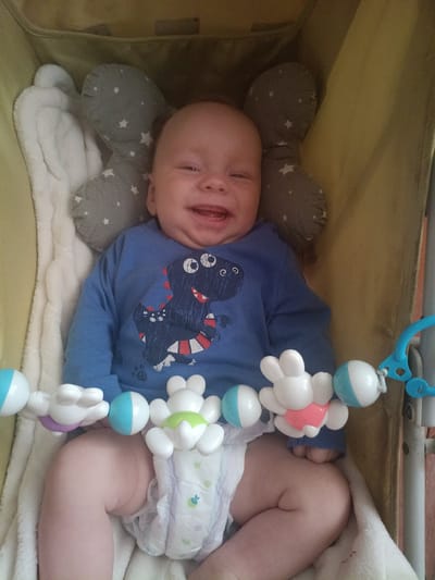 A baby lies in a stroller, facing the camera and smiling. The baby is wearing a blue long-sleeved shirt with a dinosaur print and a diaper. A mobile hangs above the baby with several colorful toys. The stroller is lined with a white blanket and a gray pillow with white stars. The background is the yellow fabric of the stroller. This image is of the city of Litynia, Ukraine.