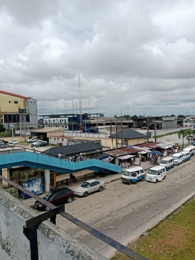 This image depicts a busy street scene in Ekurede-itsekiri, Nigeria, under a cloudy sky. In the foreground, a concrete barrier with a metal railing overlooks a road where several vehicles are parked and moving. A blue pedestrian bridge arches over the road, with a few people visible walking on it.
The road itself is lined with market stalls, covered with corrugated iron roofs, suggesting a vibrant commercial area. Various types of vehicles, including white minibuses, a dark station wagon, and a light-colored sedan, are present on the road. Some of the minibuses appear to be waiting for passengers, indicating a transport hub.
In the background, a large, multi-story building with a golden facade dominates the skyline, suggesting a modern commercial or administrative center. Several other buildings of varying sizes and architectural styles are visible, along with numerous telecommunication towers. The overall impression is one of a lively urban environment with a mix of commercial activity, transportation, and infrastructure.