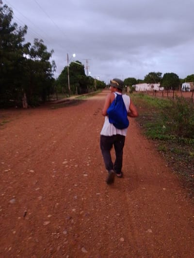 A person is depicted from behind, walking along a reddish-brown dirt road under an overcast sky, likely at dusk or late evening. The individual wears a white sleeveless shirt, dark pants, and a dark cap with a lighter embellishment, and carries a vibrant blue drawstring bag on their back. They are wearing sandals or flip-flops.

The unpaved road, scattered with small stones and gravel, stretches into the distance. Lush green vegetation, including trees and bushes, lines both sides of the road. In the background, utility poles with power lines are visible, with some streetlights illuminated, further indicating the dimming light. Beyond the foliage and fences made of wooden posts and wire, several small, simple white-walled buildings with dark roofs can be seen. The overall setting suggests a rural or semi-rural landscape in El Samán, Venezuela. No text is visible in the image. The atmosphere appears quiet and serene.