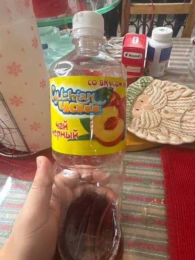An indoor scene in Matveevskoe, Russia, captures a close-up view of a table surface with various objects. A right hand, with light skin tone, holds a clear plastic bottle containing a dark brown liquid, approximately one-quarter full. The bottle features a yellow label with Cyrillic text, reading "СУЛТАН ЧАЙ" (Sultan Tea), "СО ВКУСОМ ПЕРСИКА" (with peach flavor), and "ЧАЙ ЧЕРНЫЙ" (Black Tea), accompanied by an illustration of peaches. The bottle is capped with a white screw-top lid. To the immediate left of the bottle, a white metal napkin holder contains white paper napkins adorned with red snowflake patterns. The table surface is covered by a striped tablecloth or mat with horizontal green and white bands, and a solid red textured mat is visible on the right. In the background, from left to right, obscured plastic containers are partially visible behind the napkin holder. Further right, a red cardboard box labeled "Komfi" is present, next to a white plastic bottle with a blue label section. A light beige ceramic plate, molded with a relief design resembling a stylized bearded face (possibly Santa Claus) with textured hair or a knitted cap, rests on the table behind the main bottle. A wooden furniture element with dark metal scrollwork is visible in the distant background.
