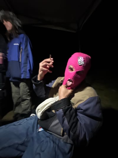 The image shows two individuals outdoors at night. One person wears a pink balaclava with a logo and holds a lit cigarette in their right hand. They wear a multi-toned jacket with blue, black, beige, and white panels, and blue pants. The other person lies down, partially visible and wearing a blue jacket. The background is dark, obscuring details but suggesting a wooded or natural environment. The presence of a tent suggests the location is a campsite. Specific location details within Auvers-Saint-Georges, France, are not discernible from the image.