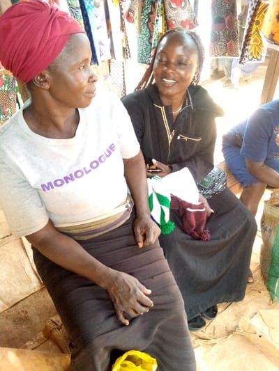 Two women are seated outside with textiles hanging in the background. The woman on the left wears a red headwrap, a grey t-shirt with the word "MONOLOGUE" printed on it, and a dark brown skirt. She looks towards the other woman. The woman on the right has braided hair and wears a black jacket with gold trim. She holds what looks like knitted scarves.