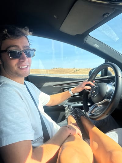 The image depicts the interior of a moving vehicle, specifically in the city of Alcanena, Portugal, under bright daylight.
A male individual is positioned in the driver's seat, smiling directly at the camera. He is a young adult with light skin and short, wavy brown hair, wearing dark rectangular sunglasses with light frames. His attire consists of a white short-sleeved t-shirt with dark text visible on the lower front, and a dark seatbelt crosses his chest. A silver-toned watch is visible on his left wrist. His right hand is placed on the black multi-function steering wheel, featuring a prominent Mercedes-Benz logo.
To the right of the driver, the lower body of a passenger is partially visible. The passenger's bare legs, light-skinned, are extended, and their right foot, adorned with a dark flat shoe featuring white polka dots, rests against a dark interior panel of the car. The driver's left hand is holding the passenger's right hand, which has a thin red string bracelet on the wrist.
The vehicle's interior is dark-toned, with visible details including a sun visor above the windshield and an "AIRBAG" label on the A-pillar. Through the front and side windows, a clear blue sky is visible above a dry, light-colored landscape featuring distant low hills with sparse vegetation, suggesting an open road or highway environment. Shadows from the bright sunlight are cast within the car's interior.