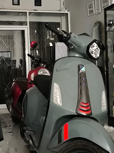 In Miruektaman, Indonesia, a close-up, eye-level shot shows two Vespas parked indoors. The foreground scooter is a muted green, its front fender decorated with a red and black stripe. Red accent lights are visible below the handlebars. Behind it, a red Vespa is partially visible. The background suggests an indoor setting with a metal security grate on a large window or door and what appears to be a retail space with shelves and displays.