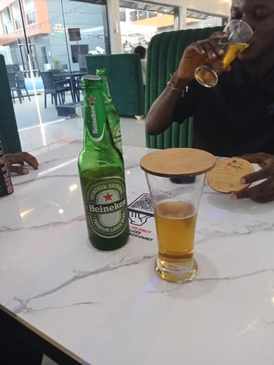 The image shows a table in a restaurant. A person is sitting in a green chair and drinking from a glass of beer. The person's arm is visible and they are wearing a black shirt. The person is holding the glass with their right hand, with a silver bracelet on their wrist. There are two bottles of Heineken beer on the table. The person is seated on the left side of the table. There is a full glass of beer on the table with a wooden coaster. There is a square, white sticker with red writing on the table. The table has a white marble surface. The background shows glass walls and a partially visible orange wall. The restaurant has a modern, clean design. There are chairs and a table visible in the background. The location is Ujevwu, Nigeria, at the coordinates 5.471 latitude and 5.818 longitude.