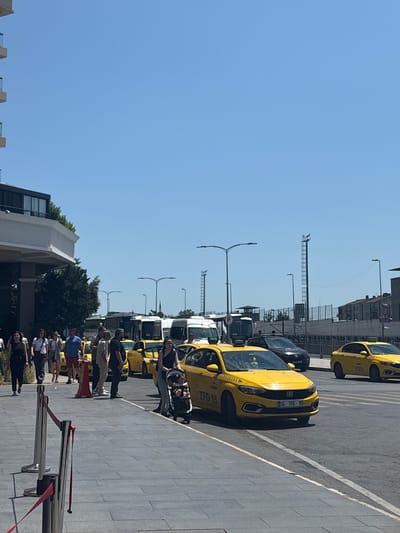 The image depicts a sunlit urban street scene in Gaziosmanpaşa, Turkey, observed from a paved sidewalk adjacent to a multi-lane road. The sky is clear and blue, indicating fair weather.
On the left, a large building with a white facade and dark upper stories featuring glass balconies is partially visible, partially obscured by green trees. In the immediate foreground on the left, a section of a gray tiled sidewalk is demarcated by chrome stanchions with red velvet ropes, suggesting a queue or restricted access area.
Numerous pedestrians are walking along this sidewalk from left to right. Among them are a woman in a black dress, a woman in a white top and dark pants, and a man in a blue t-shirt and red shorts. Further right on the sidewalk, a woman pushes a black stroller containing a child dressed in orange or red clothing. A red traffic cone stands on the sidewalk near a group of pedestrians.
The multi-lane road parallel to the sidewalk features several bright yellow taxis, consistent with Turkish taxi services. The taxi closest to the camera is a Fiat model with the license plate "34 TFG 15". It has a "TAKSI" sign on its roof. Behind it, another yellow taxi is partially visible, followed by a dark-colored sedan or SUV. Further down the road, a line of white vans or minibuses is stopped or parked, with several more yellow taxis interspersed. One distant yellow taxi on the right side of the road has a partially visible license plate "34 TTZ 59". Lane markings, including white dashed lines and a pedestrian crossing, are visible on the asphalt.
In the mid-ground and background, tall streetlights with green poles are prominent. Metal towers with ladders are also visible. A fence runs along a raised embankment in the background, behind which residential buildings with tiled roofs can be seen. A slender minaret or spire is faintly visible in the far background, rising above the white vans, confirming the Turkish setting.
