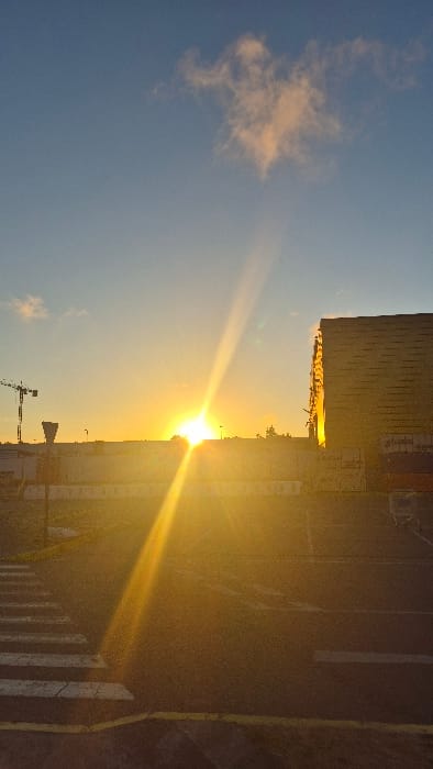 A building's facade meets a vibrant sky at sunset. Golden light emanates from the sun, casting a warm glow over the horizon. A lone cloud floats in the blue expanse. Below, a construction crane stands silhouetted against the radiant sky. A pedestrian crossing marks a path on a dark road.