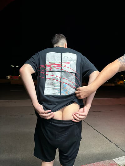 The image shows two people outdoors at night. One person, seen from behind, is wearing a black t-shirt with a graphic design and black shorts. Their t-shirt is being lifted by the second person, revealing a portion of their buttocks. The second person's arm and hand, with visible tattoos on the arm, are visible reaching under the t-shirt. The background consists of a dark, blurry area that suggests it is nighttime, with some faint lights in the distance. The ground is paved and appears to be a sidewalk or street. There is a red and white striped curb visible on the right side of the frame. The overall impression is of a casual, possibly staged, interaction between the two individuals. No text is clearly visible in the image.
